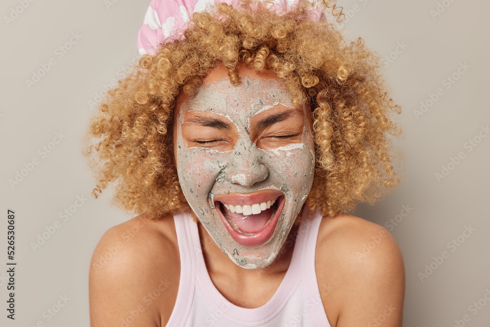 Headshot of positive curly haired woman applies beauty mud mask for reducing fine lines giggles happily dressed in t shirt stands bare shoulders isolated over grey background. Skin treatment Headshot of positive curly haired woman applies beauty mud mask for reducing fine lines giggles happily dressed in t shirt stands bare shoulders isolated over grey background. Skin treatment