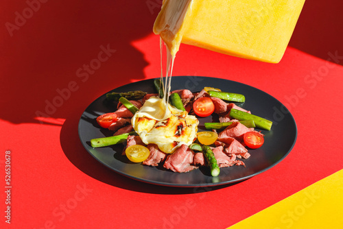 Delicious raclette cheese served over Roast Beef with Tomatoes and Asparagus, dish on red background. Hard light, deep shadow