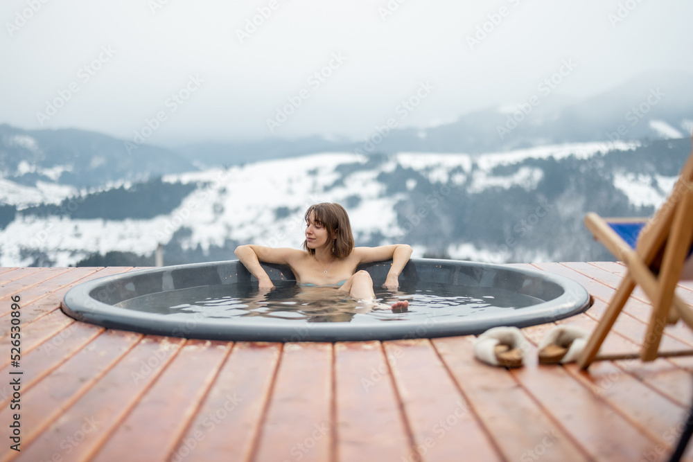 Naklejka premium Young woman bathing in hot tub at mountains during winter. Concept of rest and recovery in hot vat on nature. Idea of escape and recreation on mountains