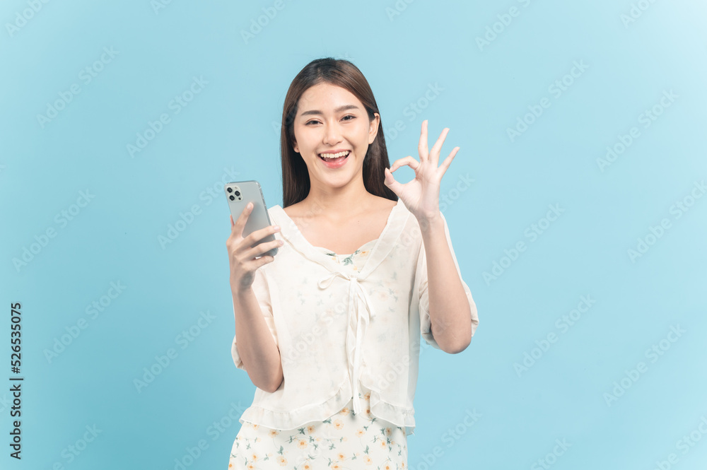Beautiful young asian woman in white dress with flower pattern using smartphone show okay sign isolated on blue background Beautiful young asian woman in white dress with flower pattern using smartphone show okay sign isolated on blue background
