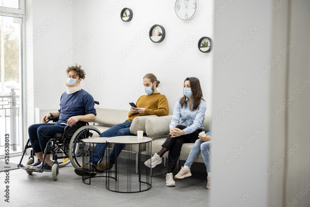 Young people in medical masks sitting in the hall of the clinic, waiting for an appointment. Man with a neck injury in a wheelchair, young adult man and woman with child on the couch