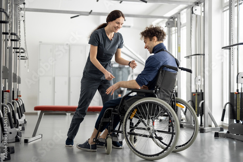 Rehabilitation specialist helps a guy stand out of a wheelchair at rehabilitation center. Concept of physical therapy and support for people with disabilities © rh2010
