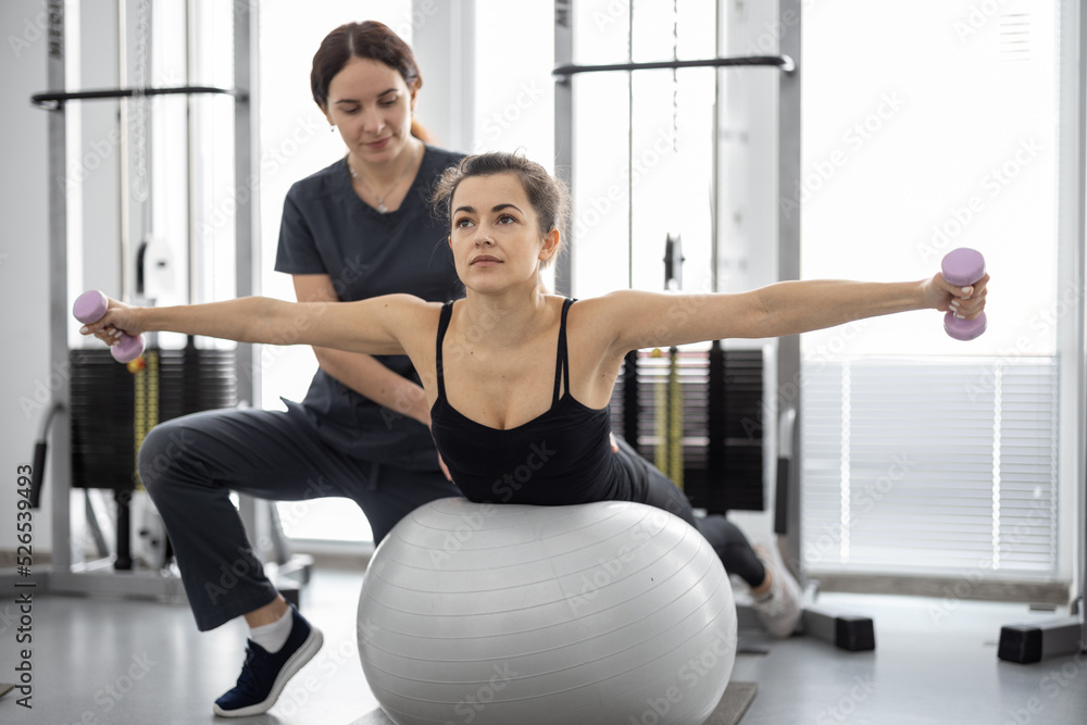 Woman doing exercises on fitness ball and dumbbells with rehabilitation specialist at the gym. Concept of physical therapy for back strengthening and recovery. Idea of recovery after pregnancy