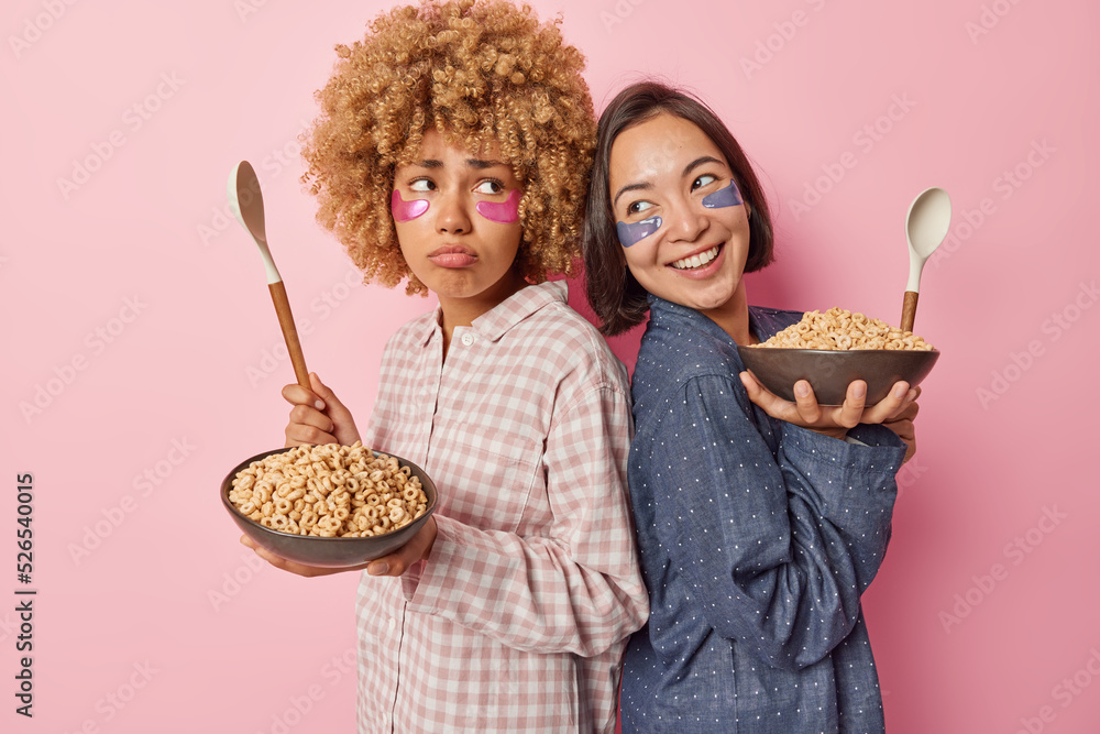 Horizontal shot of two mixed race women stand backs to each other apply beauty patches under eyes dressed in pajamas eat cereals for breakfast isolated over pink background. Morning routines Horizontal shot of two mixed race women stand backs to each other apply beauty patches under eyes dressed in pajamas eat cereals for breakfast isolated over pink background. Morning routines