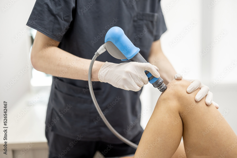 Doctor applies shock wave therapy with special medical equipment on women's knee joints at medical office. Concept of non-invasive technology for treating pain in musculoskeletal system