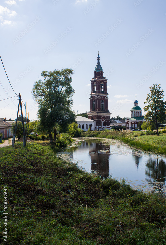 The bell tower of the Intercession Church on the river bank with reflection in the water and lush green foliage of trees on a bright sunny summer day in Yuriev-Polsky russia