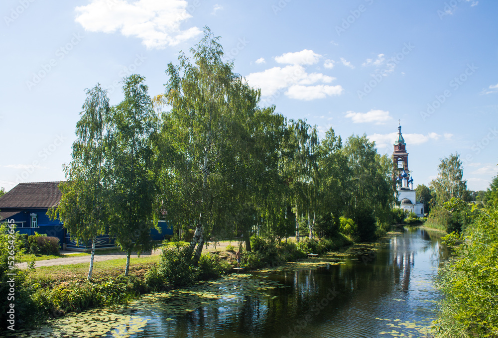 The bell tower of the Intercession Church on the river bank with reflection in the water and lush green foliage of trees on a bright sunny summer day in Yuriev-Polsky russia