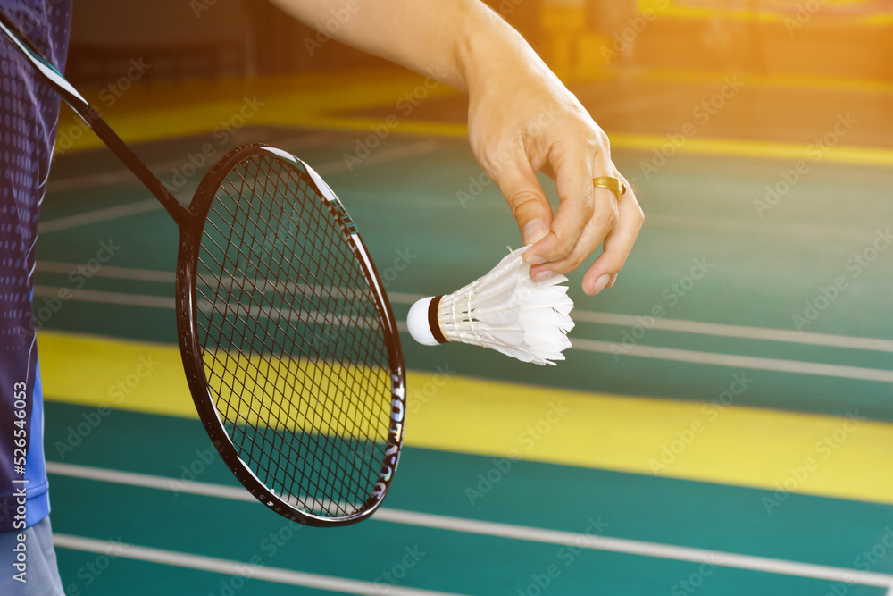 Badminton racket and old white shuttlecock holding in hands of player while serving it over the net ahead, blur badminton court background and selective focus