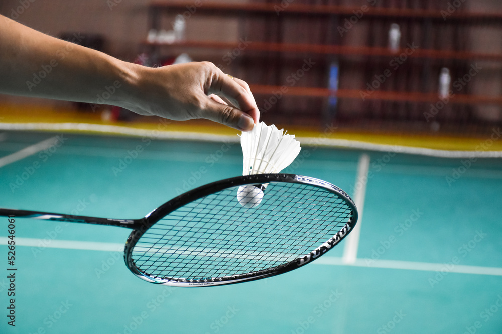 Badminton racket and old white shuttlecock holding in hands of player while serving it over the net ahead, blur badminton court background and selective focus