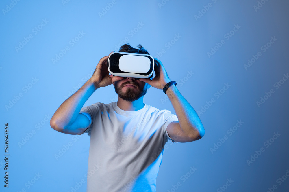 Bearded man in a white t-shirt wearing VR glasses and speaking with a friends in metaverse in a room with blue light. VR helmet for entertainment, learning and gaming. Bearded man in a white t-shirt wearing VR glasses and speaking with a friends in metaverse in a room with blue light. VR helmet for entertainment, learning and gaming.