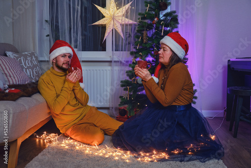 An adult man and a woman drink from a cup at the Christmas tree. Home family comfort on New Year Eve in the evening living room