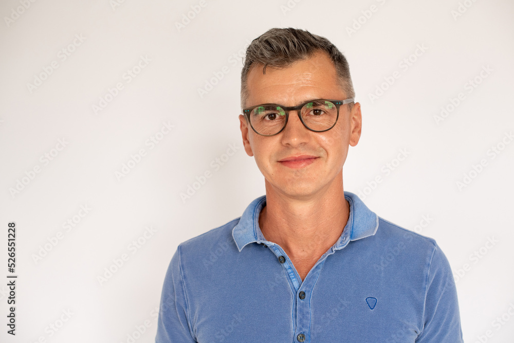 Portrait of mature man wearing glasses smiling at camera. Caucasian man wearing blue T-shirt standing and looking at camera against white background. Eyesight concept Portrait of mature man wearing glasses smiling at camera. Caucasian man wearing blue T-shirt standing and looking at camera against white background. Eyesight concept