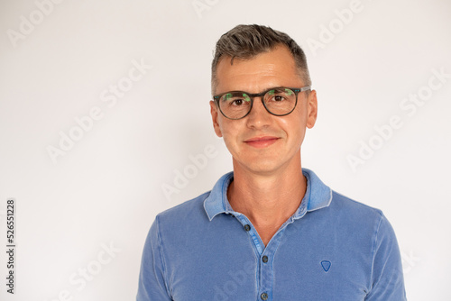 Portrait of mature man wearing glasses smiling at camera. Caucasian man wearing blue T-shirt standing and looking at camera against white background. Eyesight concept