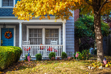 Blue shinngled two stor house entrance with porch and rocking chairs and American flags and a fall wreath under a yellow-leafed Autumn maple tree - closeup