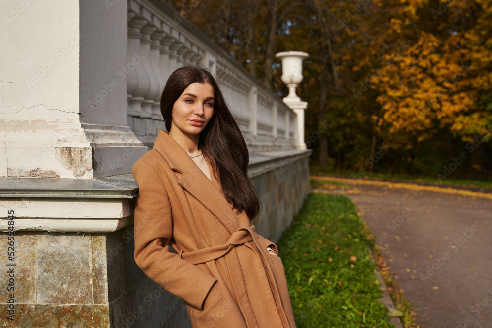 Naklejka premium a beautiful woman in a brown coat smiles and stands leaning against white columns in an autumn park