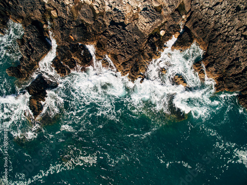 Small waves repeatedly crash on a small sandy beach in a bay with an uneven rocky coastline, aerial photography overhead.