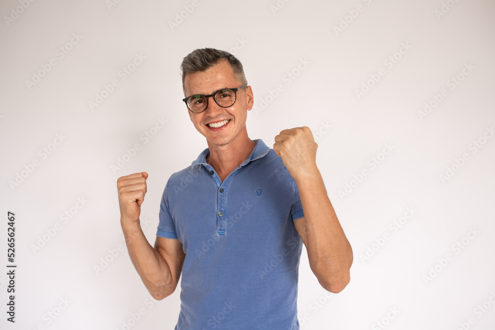 Portrait of happy mature man making winning gesture. Caucasian man wearing blue T-shirt and eyeglasses smiling at camera and showing fists over white background. Success concept Portrait of happy mature man making winning gesture. Caucasian man wearing blue T-shirt and eyeglasses smiling at camera and showing fists over white background. Success concept