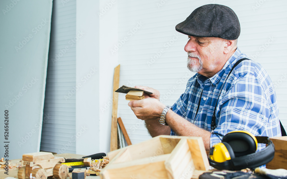Senior Caucasian happy retired old male woodworker or carpenter smiling, wearing check shirt with apron, vintage hat, creating DIY wooden mini figure toy for decoration as hobby after retirement. Senior Caucasian happy retired old male woodworker or carpenter smiling, wearing check shirt with apron, vintage hat, creating DIY wooden mini figure toy for decoration as hobby after retirement.