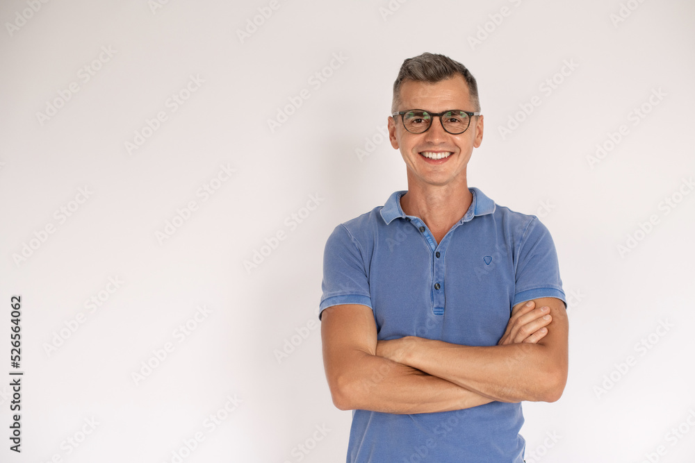 Portrait of smiling mature man standing with folded arms. Happy Caucasian man wearing blue T-shirt and eyeglasses standing over white background. Eyesight concept Portrait of smiling mature man standing with folded arms. Happy Caucasian man wearing blue T-shirt and eyeglasses standing over white background. Eyesight concept