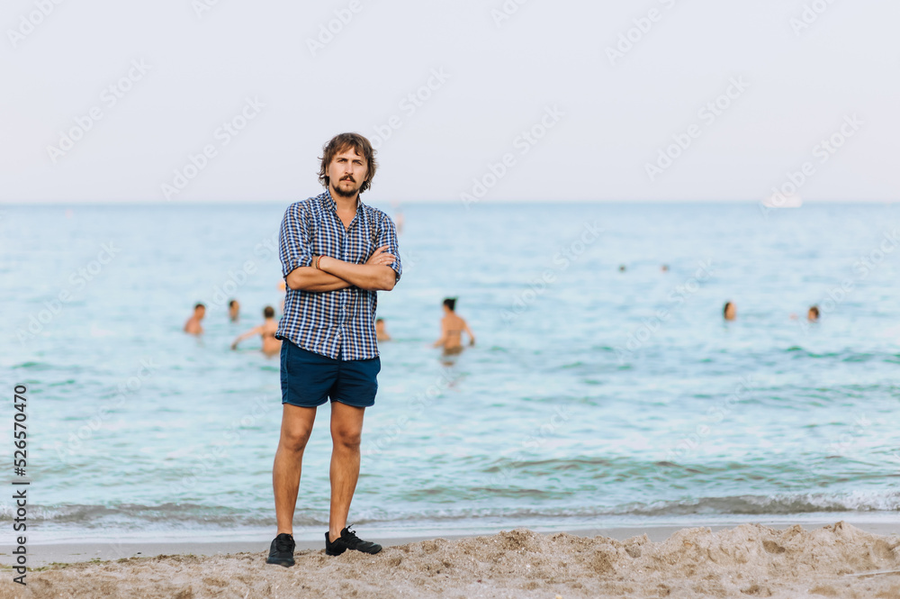 A handsome, young hippie man in a shirt and shorts stands on the beach on the sand against the backdrop of the sea, ocean and people swimming in the summer season.
