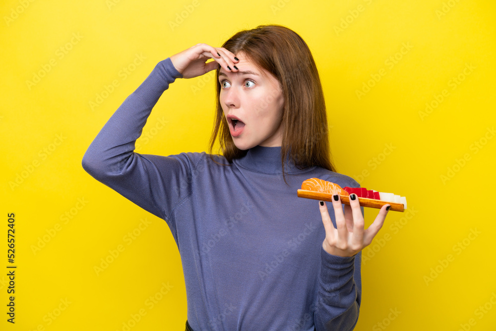 Young English woman holding sashimi isolated on yellow background doing surprise gesture while looking to the side Young English woman holding sashimi isolated on yellow background doing surprise gesture while looking to the side