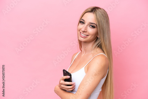 Pretty blonde woman isolated on pink background holding a mobile phone and with arms crossed