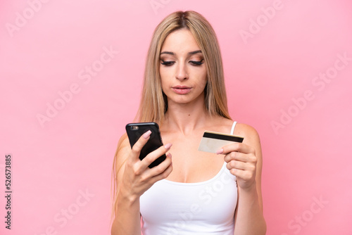 Pretty blonde woman isolated on pink background buying with the mobile with a credit card