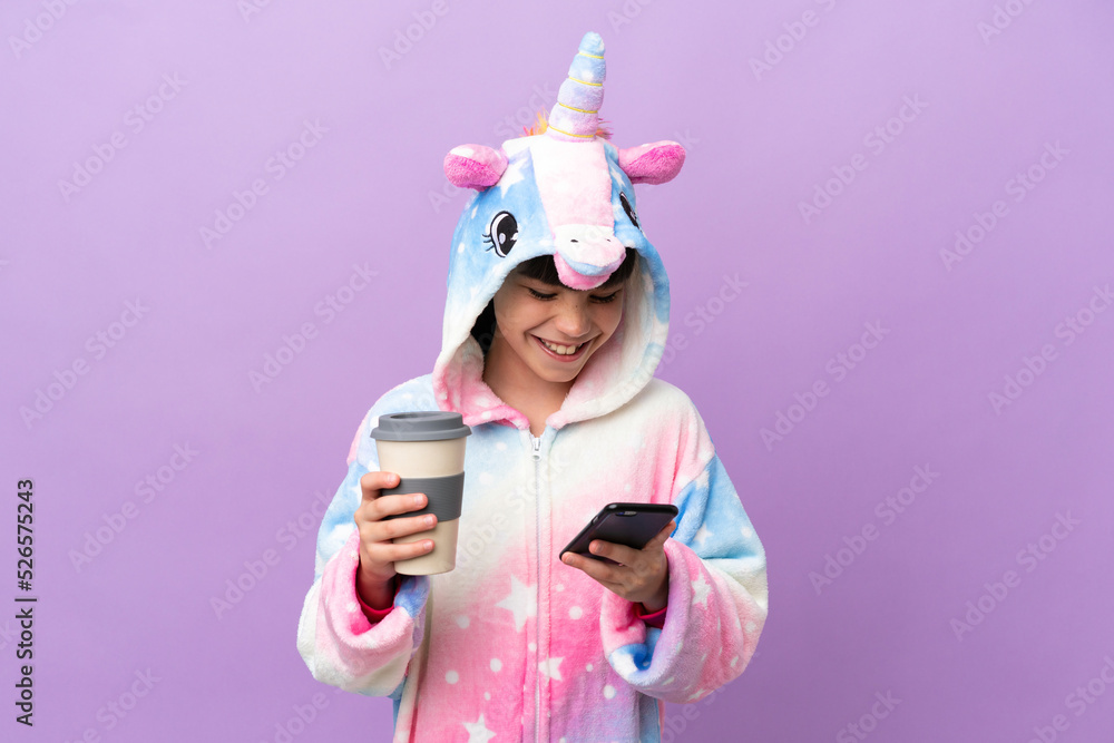 Little kid wearing a unicorn pajama isolated on purple background holding coffee to take away and a mobile Little kid wearing a unicorn pajama isolated on purple background holding coffee to take away and a mobile