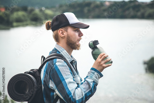 A man traveler with a backpack and a karemat keeps drinking water from a flask.