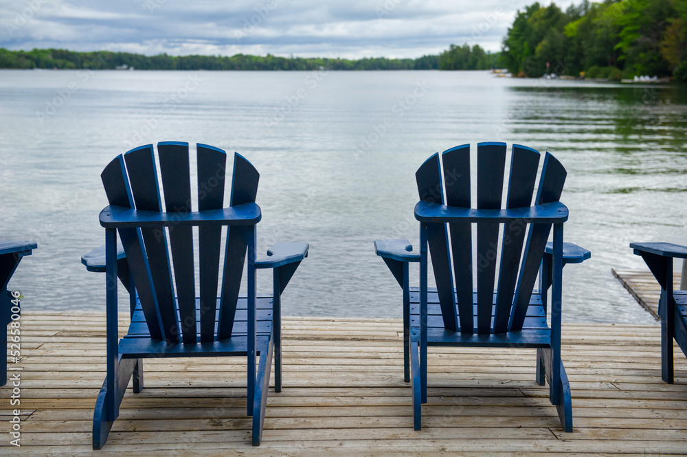 Fototapeta premium Close up of two blue Adirondack chairs on a wooden dock facing a lake in Muskoka, Ontario.