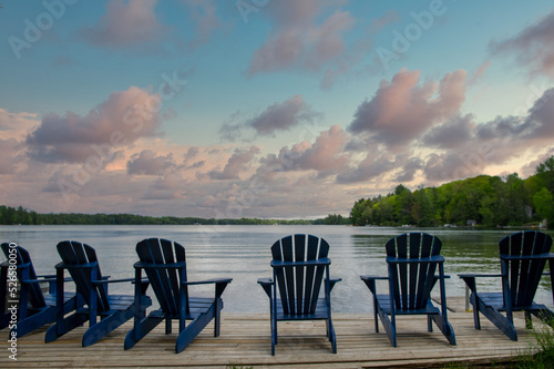 Multiple blue Adirondack chairs on a wooden dock facing a lake in Muskoka, Ontario, at sunset. Beautiful clouds are visible in the sky.