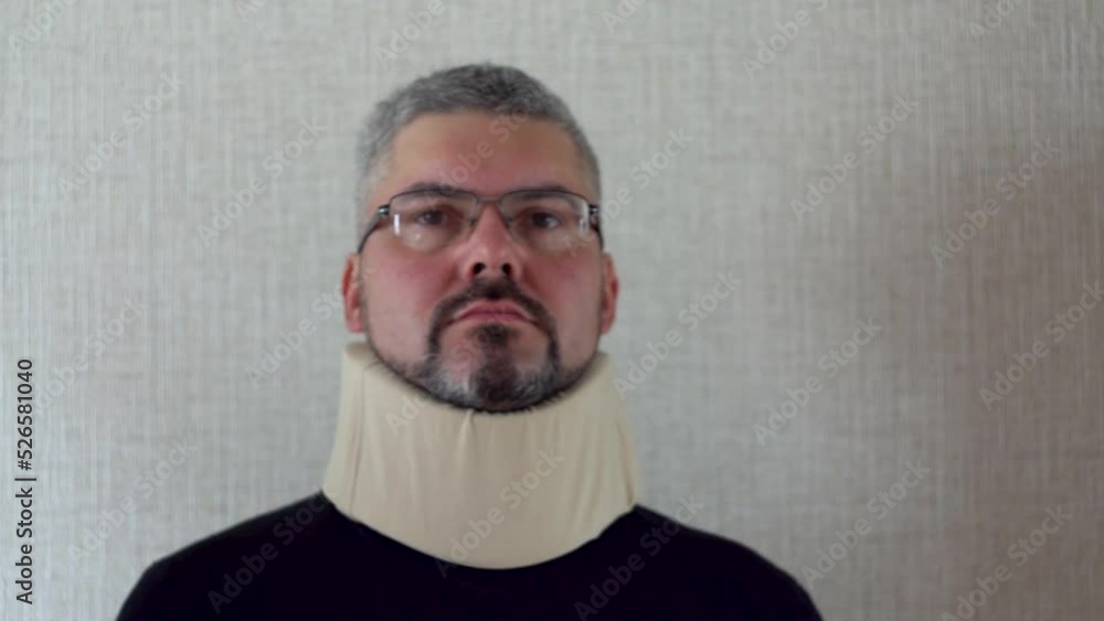 An adult man is standing against a wall. An orthopedic collar is worn