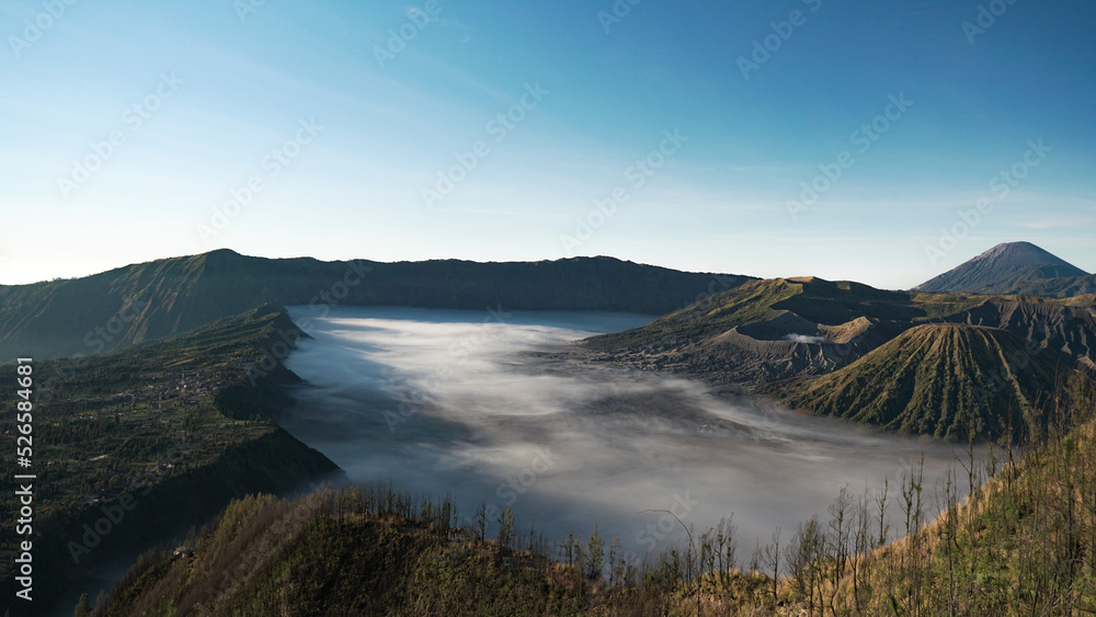 mystic and surreal panoramic mountain view of Bromo volcano craters with foggy surface and green vegetation including Cemoro Lawang village during morning sunshine
