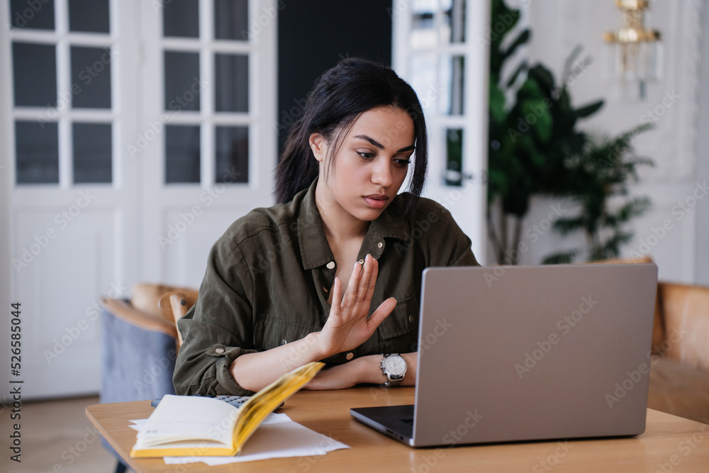 Stop it! Tired young Brazilian office manager making stop gesture by palm during video call, sitting at table with laptop. Upset woman entrepreneur refusing to continue partnership. Business people. Stop it! Tired young Brazilian office manager making stop gesture by palm during video call, sitting at table with laptop. Upset woman entrepreneur refusing to continue partnership. Business people.