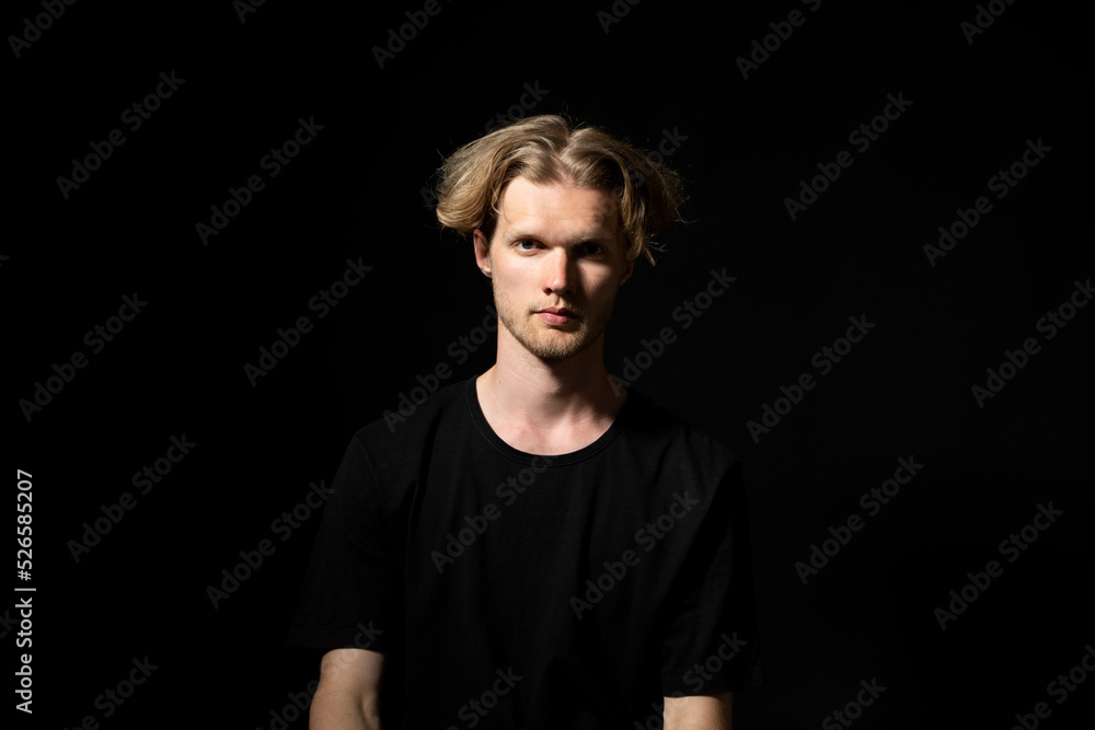Naklejka premium Portrait of calm serious mature young man without emotions. Beautiful emotionless guy in a black t-shirt looking to the camera, isolated over black background.