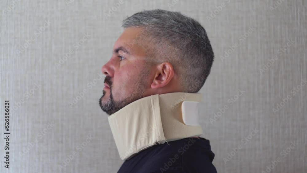 An adult man is standing against a wall. An orthopedic collar is worn