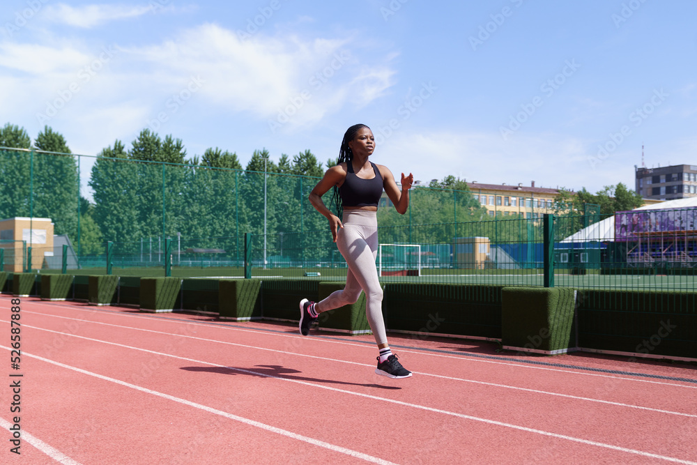 Young African American athlete with long braids runs on red track of city stadium preparing for competitions. Fit black woman in sportswear trains hard to win upcoming marathon on hot summer day
