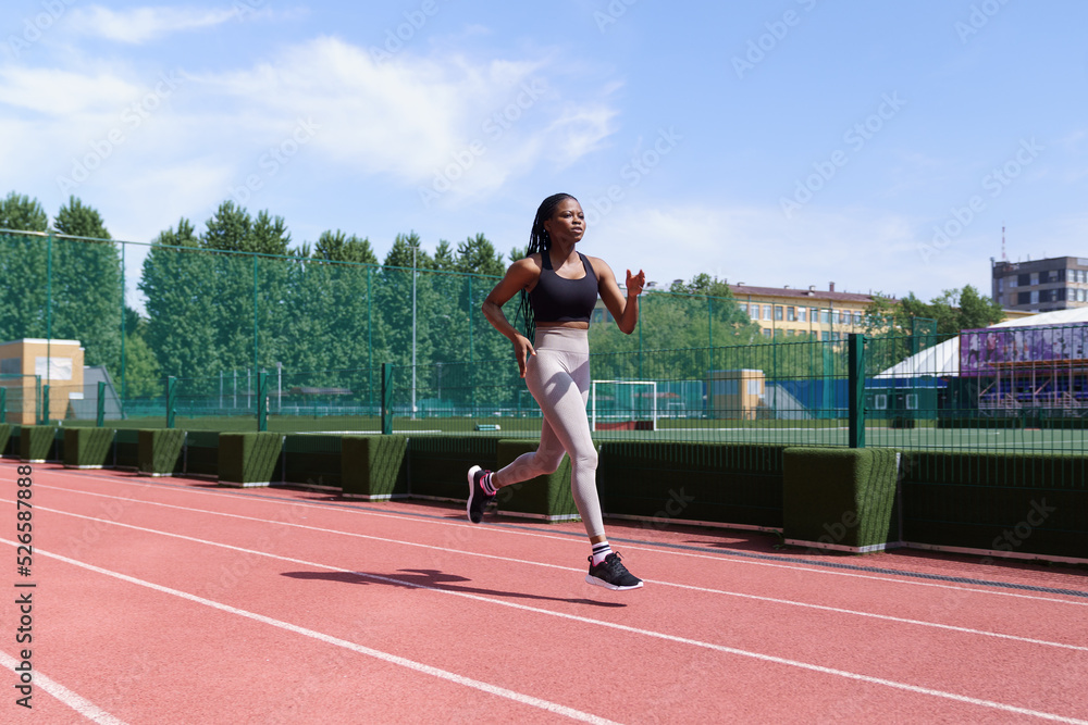 Obraz premium Young African American athlete with long braids runs on red track of city stadium preparing for competitions. Fit black woman in sportswear trains hard to win upcoming marathon on hot summer day