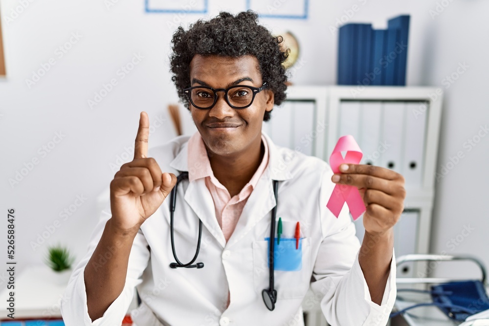 African doctor man holding pink cancer ribbon surprised with an idea or question pointing finger with happy face, number one African doctor man holding pink cancer ribbon surprised with an idea or question pointing finger with happy face, number one