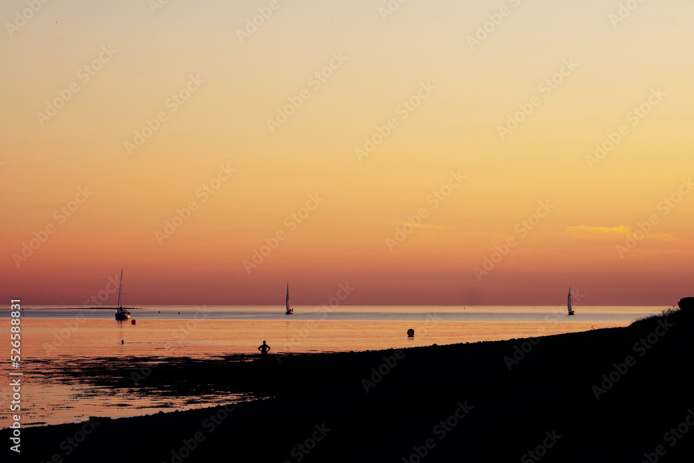 Sunrise scene by the ocean with warm orange color. Silhouette of a small yachts sailing. Calm and peaceful scene.
