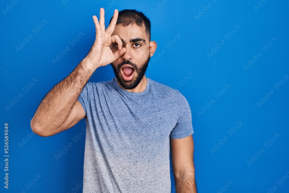 Middle east man with beard standing over blue background doing ok gesture shocked with surprised face, eye looking through fingers. unbelieving expression.