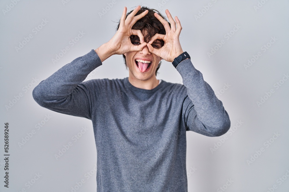Young man standing over isolated background doing ok gesture like binoculars sticking tongue out, eyes looking through fingers. crazy expression. Young man standing over isolated background doing ok gesture like binoculars sticking tongue out, eyes looking through fingers. crazy expression.