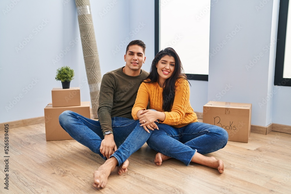 Young couple sitting on the floor at new home winking looking at the camera with sexy expression, cheerful and happy face.
