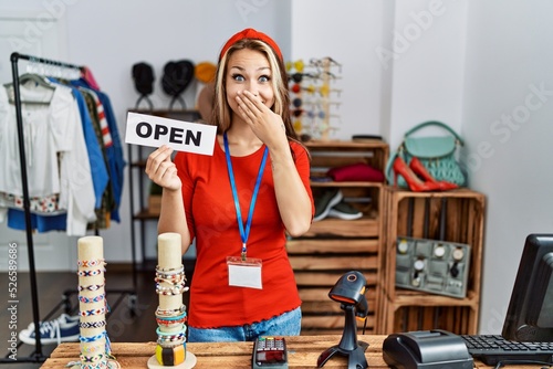 Young caucasian woman holding banner with open text at retail shop covering mouth with hand, shocked and afraid for mistake. surprised expression