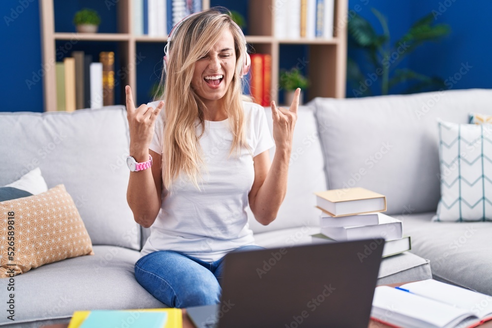 Young blonde woman studying using computer laptop at home shouting with crazy expression doing rock symbol with hands up. music star. heavy music concept. Young blonde woman studying using computer laptop at home shouting with crazy expression doing rock symbol with hands up. music star. heavy music concept.