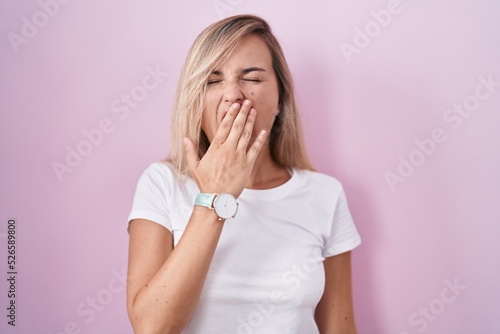 Young blonde woman standing over pink background bored yawning tired covering mouth with hand. restless and sleepiness.