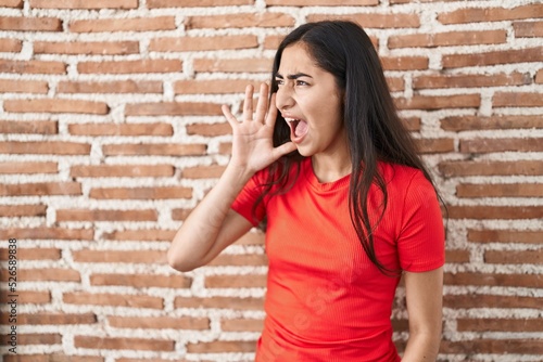 Young teenager girl standing over bricks wall shouting and screaming loud to side with hand on mouth. communication concept.