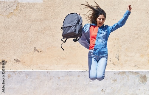 Young hispanic girl student holding backpack jumping at street