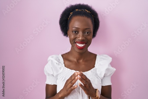 African woman with curly hair standing over pink background hands together and fingers crossed smiling relaxed and cheerful. success and optimistic