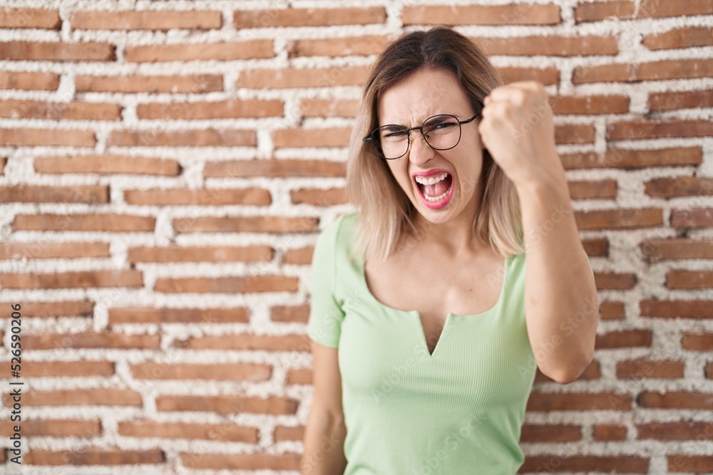 Young beautiful woman standing over bricks wall angry and mad raising fist frustrated and furious while shouting with anger. rage and aggressive concept.